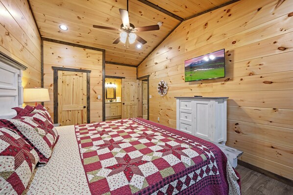 Una habitación de cabaña de madera con una cama, un ventilador de techo, un televisor montado en la pared y una puerta que da a otra habitación.