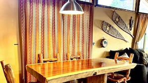 Une table à manger en bois avec des chaises, un luminaire suspendu et un rideau à motifs dans une pièce chaleureuse.