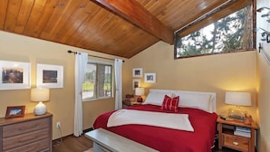 Une chambre avec un plafond en bois, un grand lit avec une literie rouge, un banc, des tables de chevet avec des lampes et des tableaux encadrés aux murs.