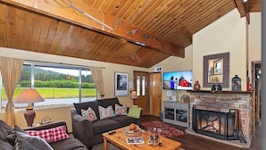 Un salon chaleureux avec un plafond en bois, une cheminée, un téléviseur et une grande fenêtre donnant sur un paysage verdoyant.