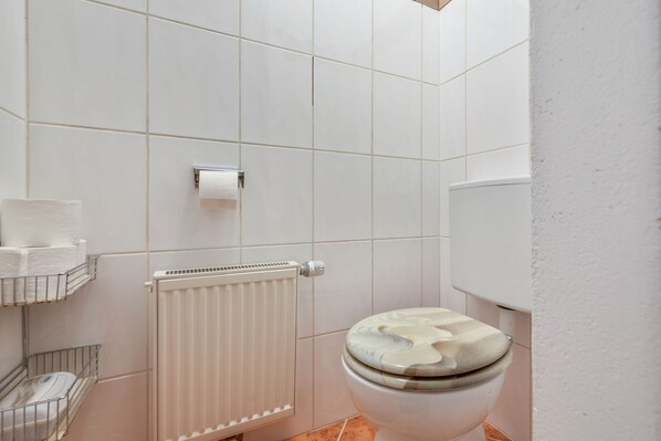 A bathroom with white tiles, a toilet, a radiator, and shelves with toiletries.