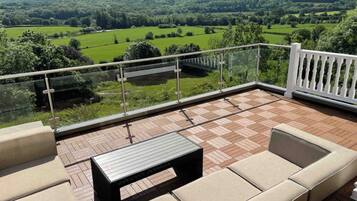 A balcony with a checkered floor, cushioned seating, and a glass railing overlooking a green landscape.