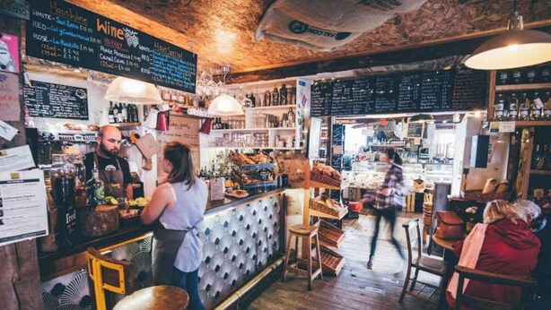 Un bar rustique avec un plancher en bois et des étagères remplies de bouteilles. Un comptoir où une personne sert des boissons, et une autre personne se tient à l’arrière-plan.