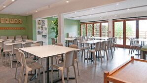 Un'ampia sala da pranzo con diversi tavoli e sedie, grandi finestre che offrono vista sul verde e una parete verde decorata con quadri.