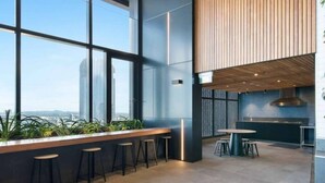 A modern interior with large windows, a bar area with stools, a kitchen, and a view of the cityscape. - 2BR Luxury Skytower CBD Pool Gym Netflix (Brisbane)