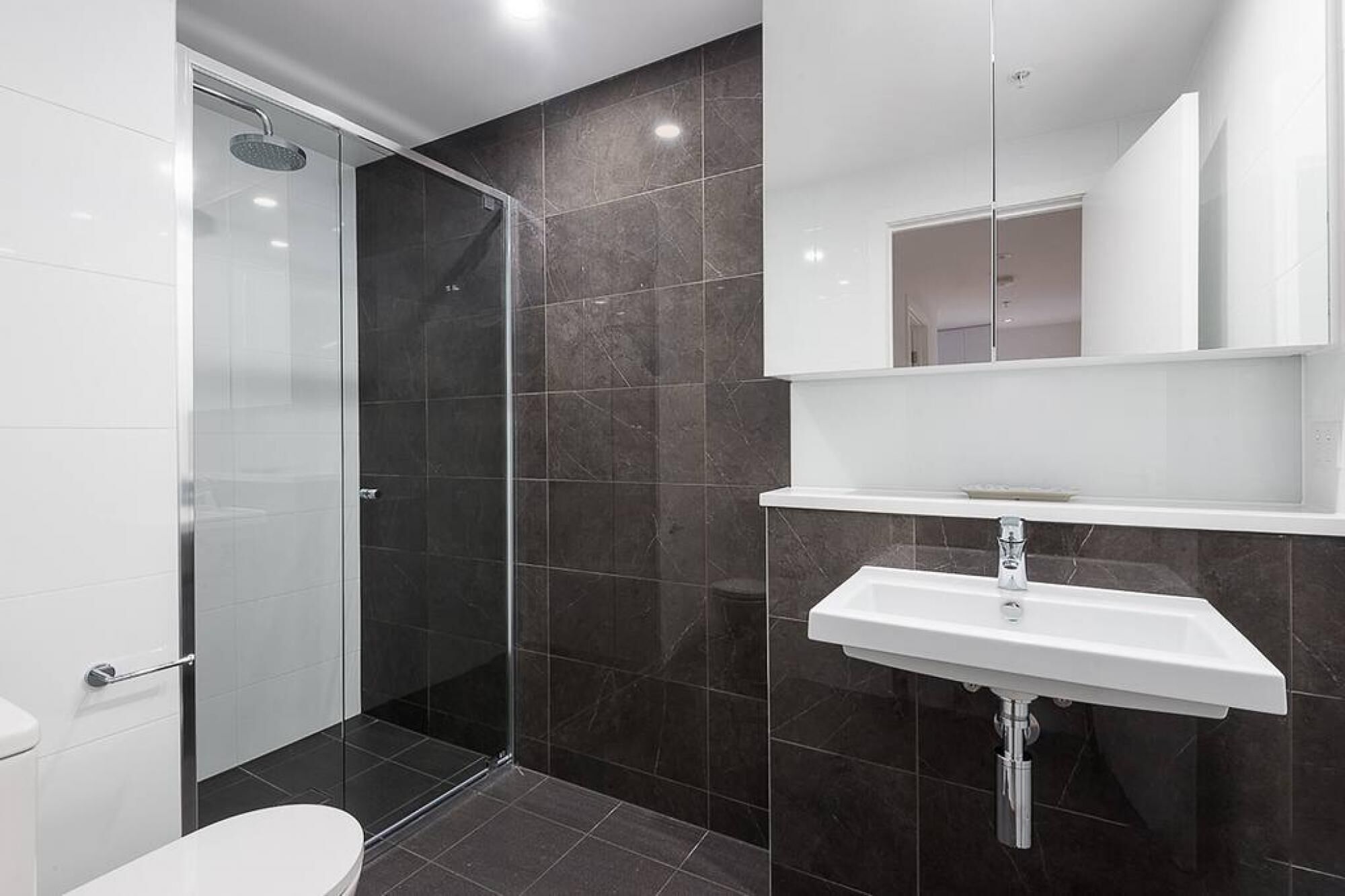 A modern bathroom with a glass-enclosed shower, a white sink, and dark tiled walls.