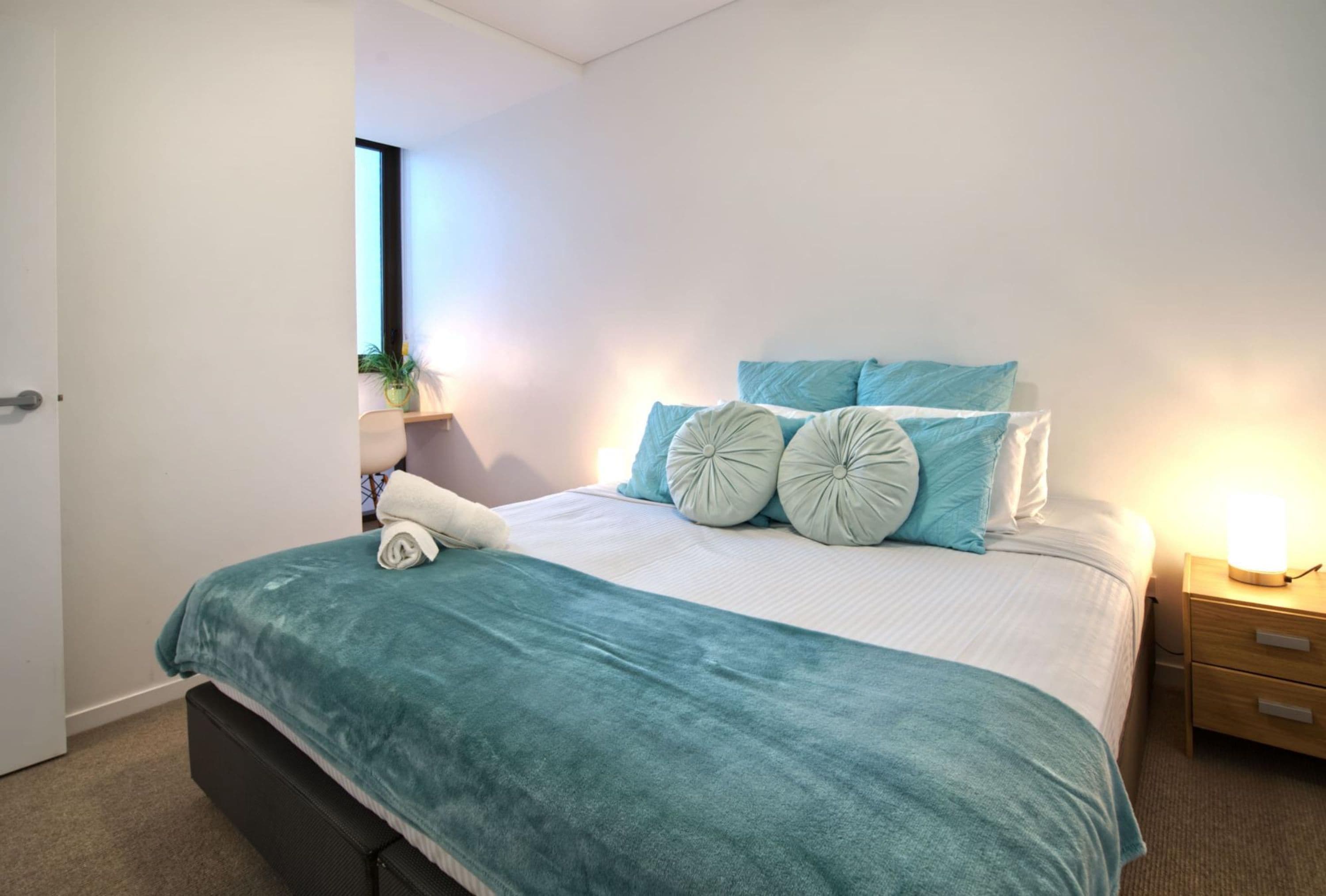 A neatly made bed with a teal bedspread and pillows, a bedside lamp, and a small shelf with a plant.