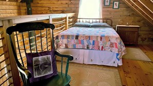 Een gezellige slaapkamer onder een schuin houten dak, met een bed met een lappendeken, een schommelstoel en een klein houten nachtkastje.