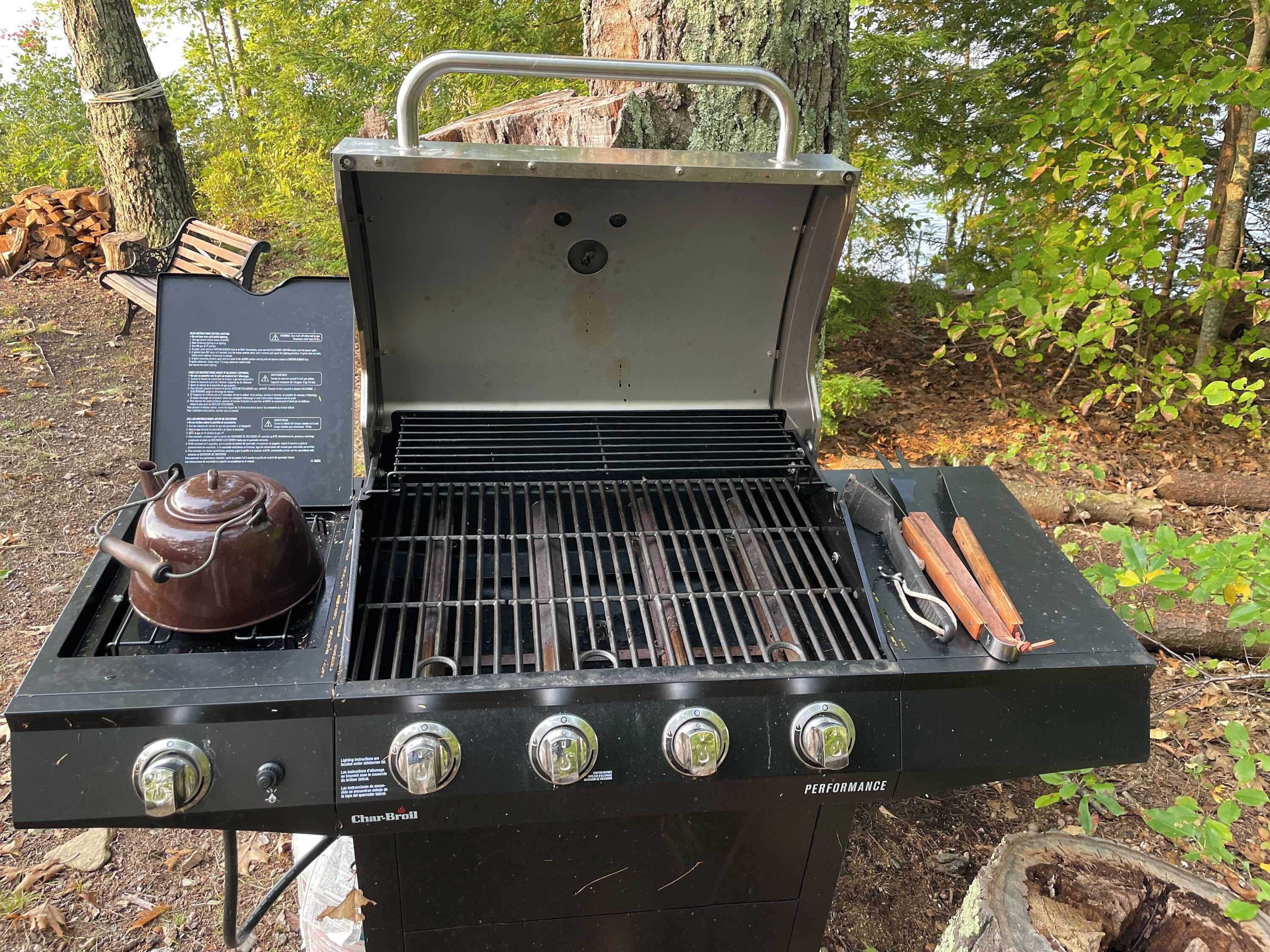 An outdoor charcoal grill with a kettle and utensils, surrounded by trees.