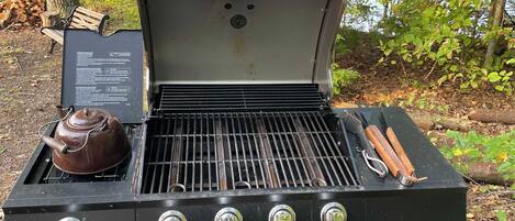 An outdoor charcoal grill with a kettle and utensils, surrounded by trees.