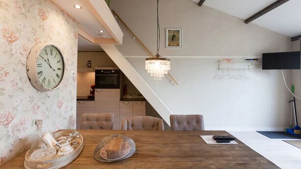 A dining area with a wooden table, cushioned chairs, a large wall clock, and a chandelier. - Nice vacation home in Horssen with garden (Horssen)
