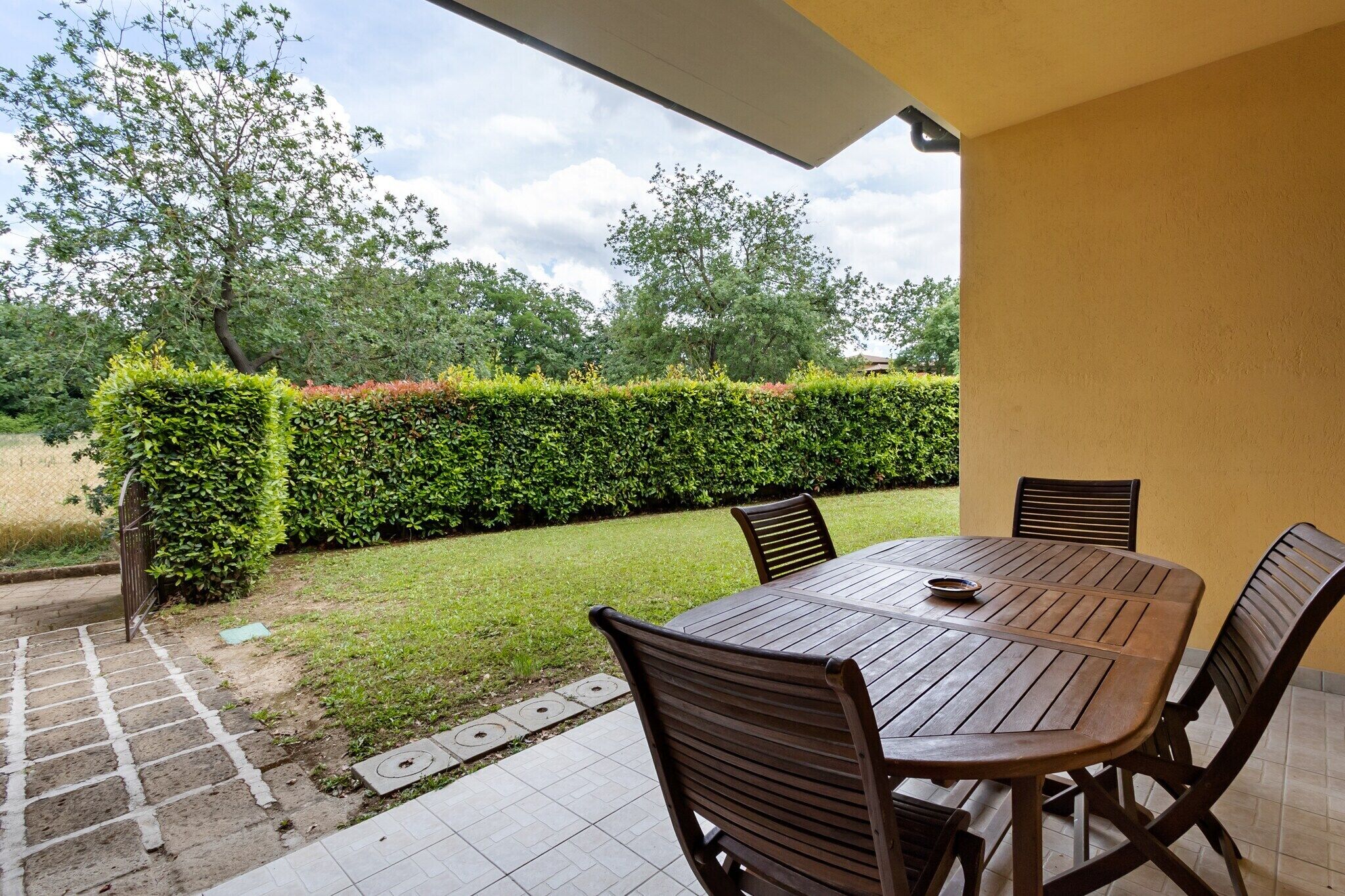 An outdoor patio with a wooden table and chairs, surrounded by a well-maintained hedge and a grassy area.