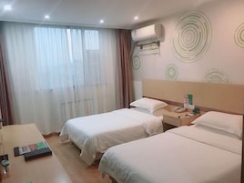 Habitación de hotel con dos camas, una mesita de noche con un teléfono, una pequeña mesa con un libro y un objeto verde, y una ventana con cortinas traslúcidas.
