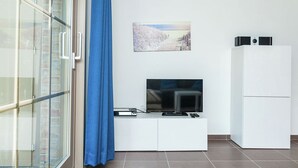 Ett modernt vardagsrum med en platt-TV placerad på ett vitt skåp, en tavla på väggen och ett vitt kylskåp.