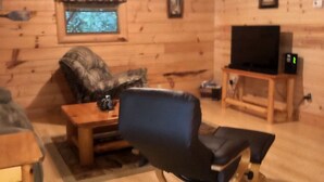 L’intérieur chaleureux d’un chalet avec un plancher en bois, un ventilateur de plafond, un t éléviseur posé sur un meuble en bois, et un coin salon confortable avec un fauteuil et un canapé.