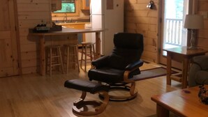 L’intérieur chaleureux d’un chalet avec un plafond en bois, un fauteuil en cuir noir, une table à manger en bois et un coin cuisine équipé d’un réfrigérateur et d’un évier.