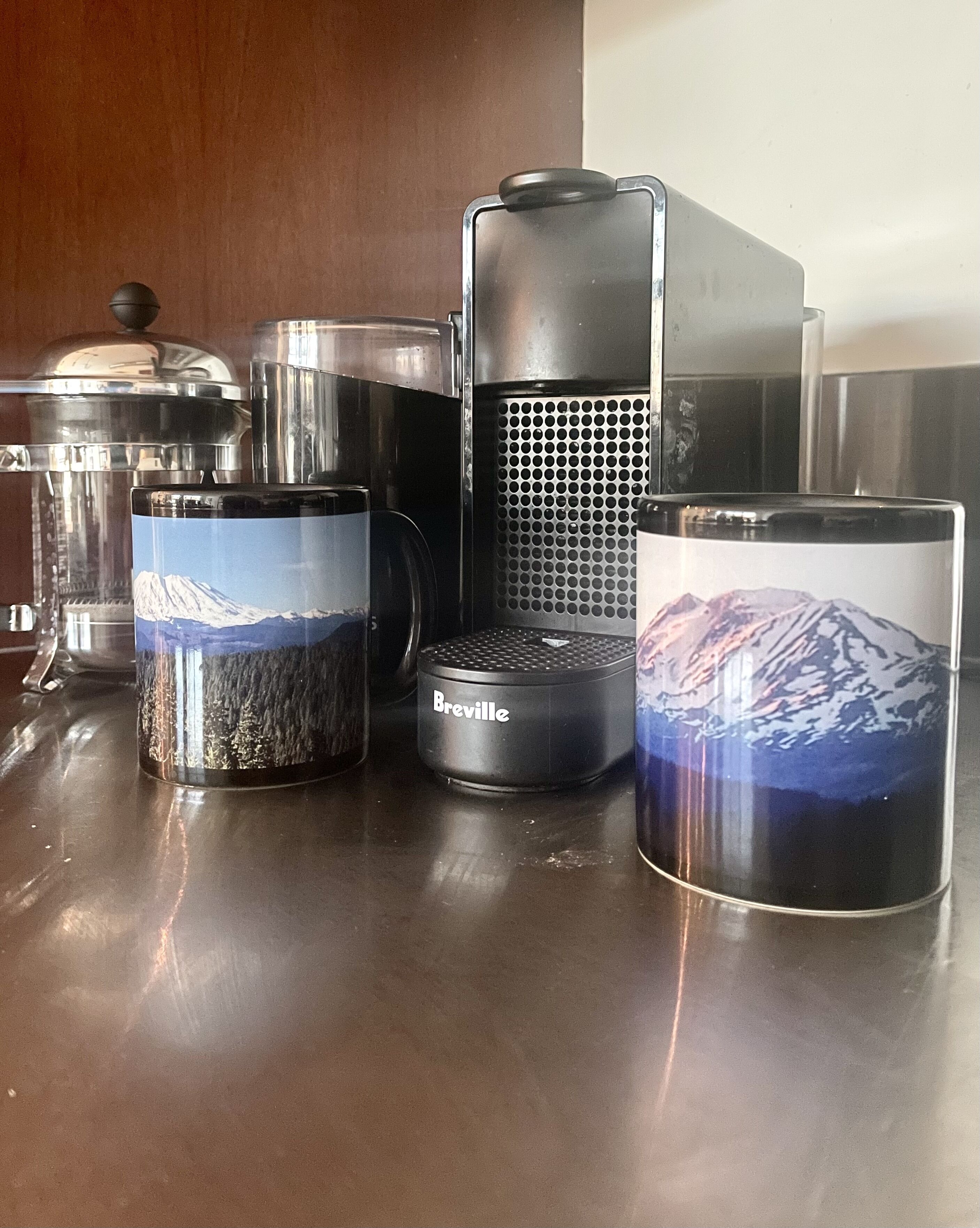 A kitchen counter with various coffee-making equipment, including a Breville coffee maker and two coffee containers with mountain images.