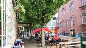 Un café en plein air avec des tables et des parasols rouges, un bâtiment en briques doté de fenêtres en arc de cercle, et un trottoir bordé d’arbres.