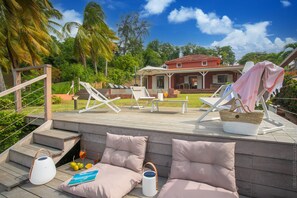 Une terrasse en bois avec des chaises longues et des coussins, un espace salon couvert et une maison en arrière-plan.