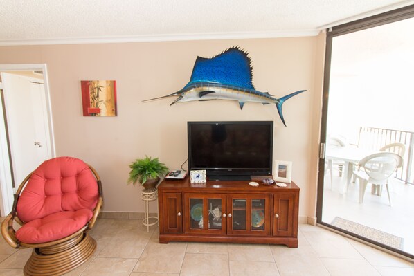 A living room with a large flat-screen TV, a red cushioned chair, a wooden cabinet with glass doors, and a decorative fish sculpture on the wall.