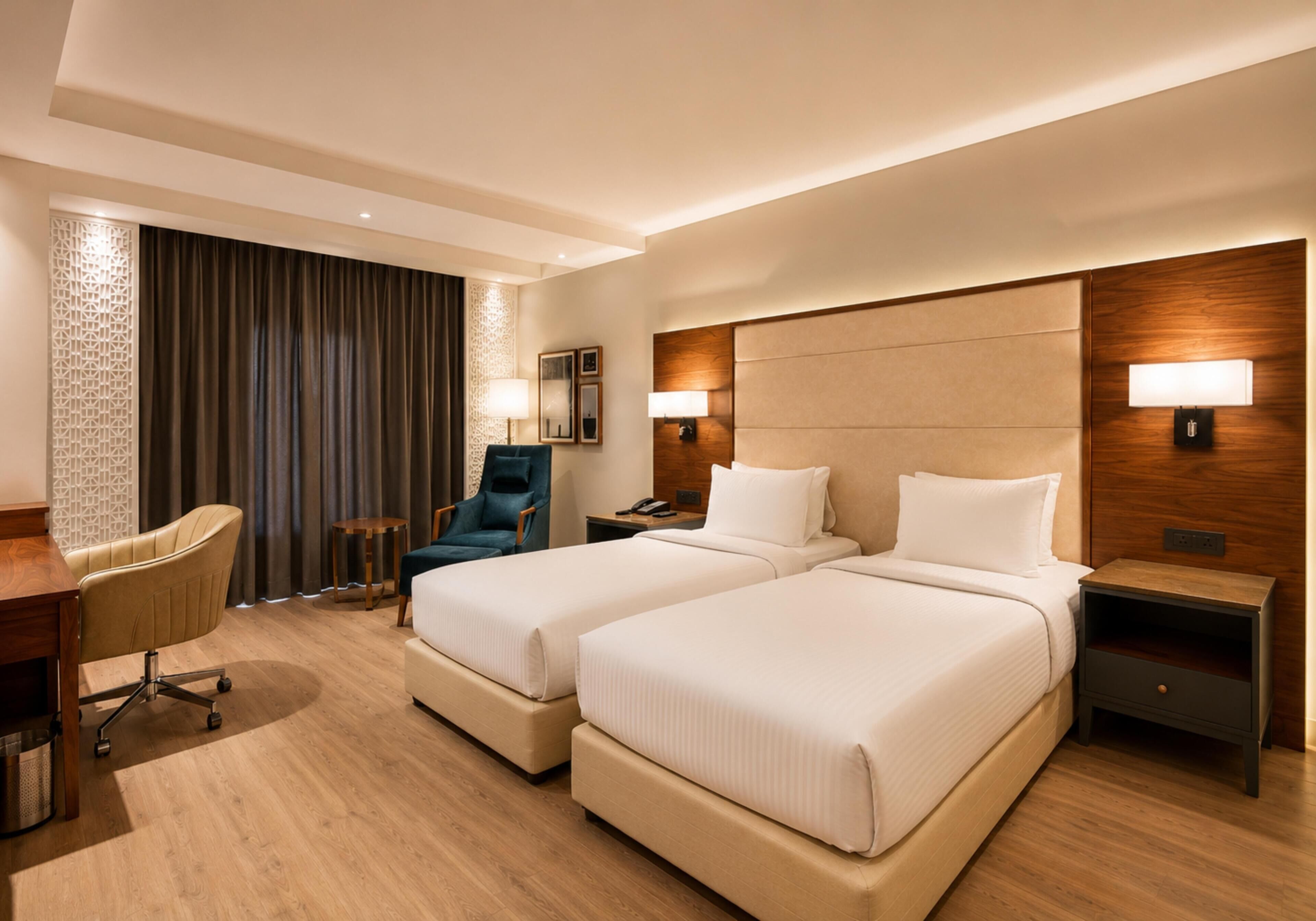 A hotel room with two beds, a desk, a chair, and a lamp.