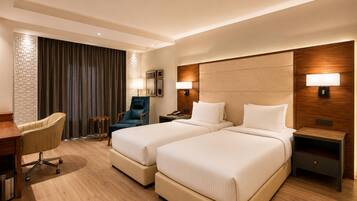 A hotel room with two beds, a desk, a chair, and a lamp.
