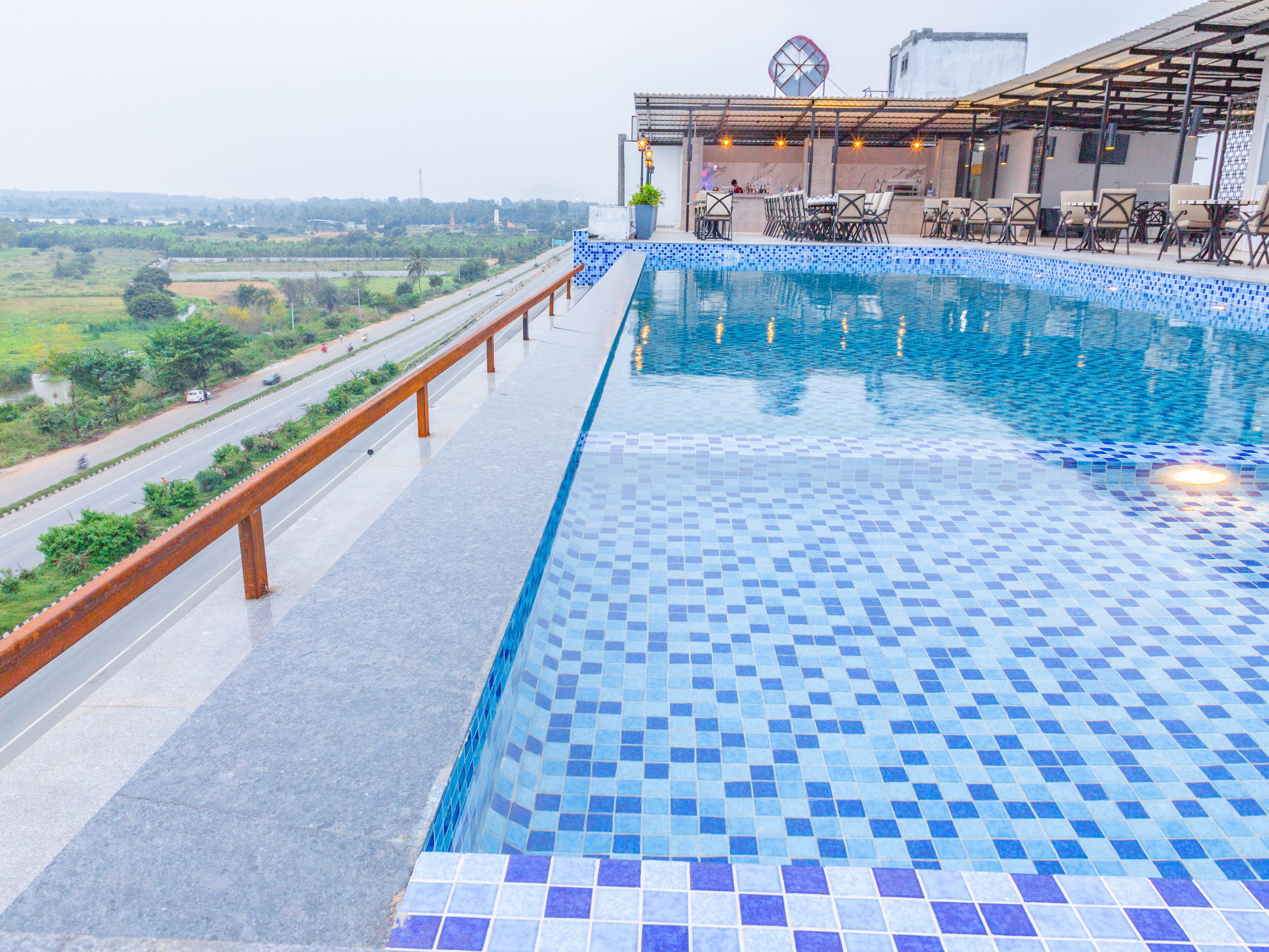 A rooftop pool with a tiled edge, a seating area with chairs, and a view of a road and greenery.