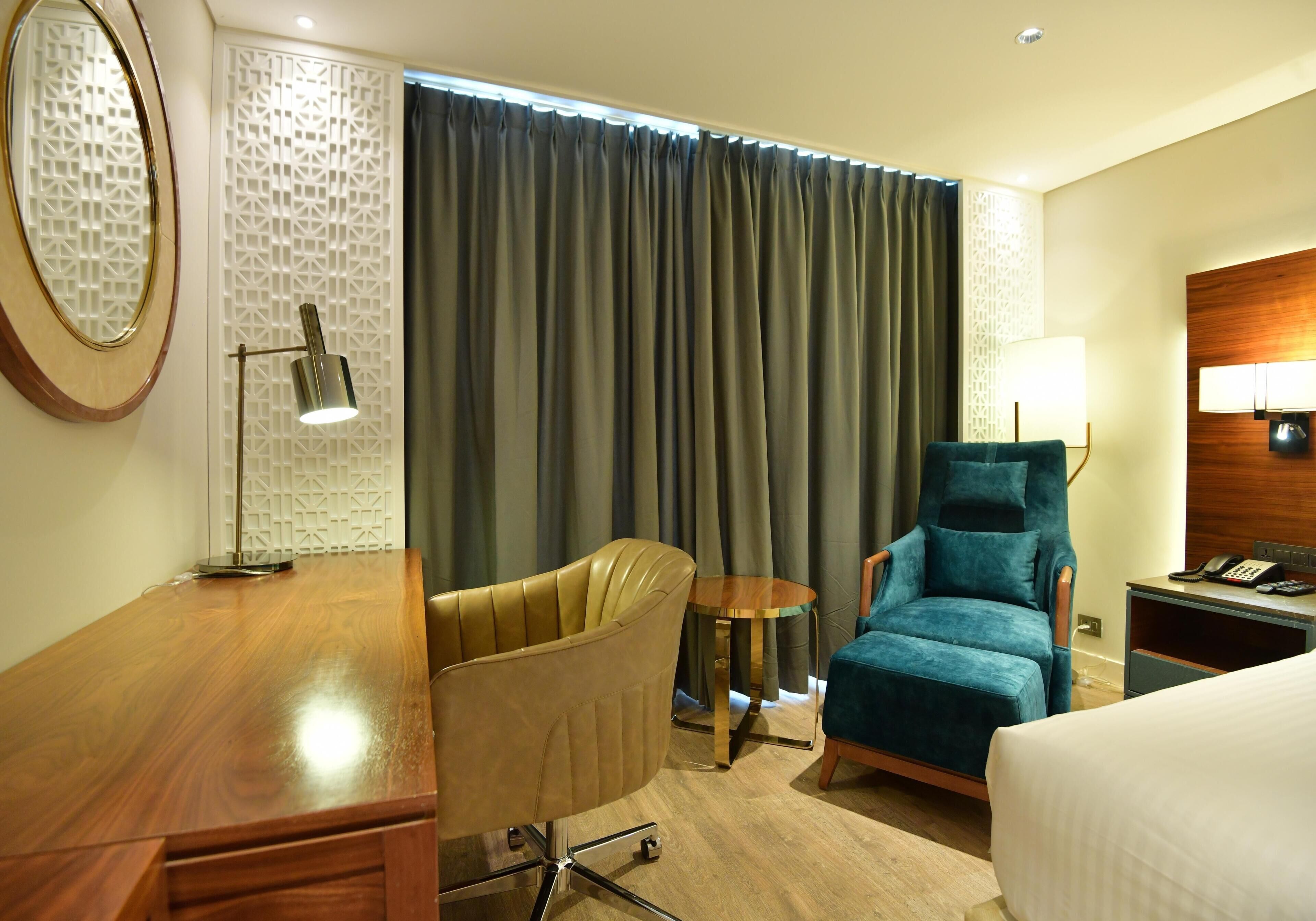 Habitación de hotel con un escritorio de madera, una silla de cuero, un sillón azul, una pequeña mesa redonda, una cama y un teléfono instalado en la pared.