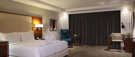 A hotel room with a large bed, a desk with a chair, a blue armchair, and a small table with a lamp.