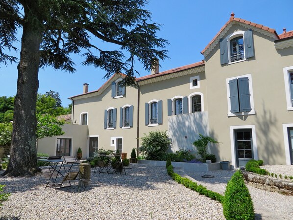 A two-story building with a symmetrical facade, multiple windows with shutters, and a gravel courtyard with a table and chairs.