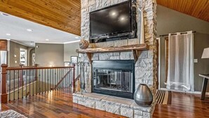Un vaste salon agrémenté d’une cheminée en pierre, d’un téléviseur à écran plat fixé au-dessus et d’un escalier en bois menant à un autre niveau.