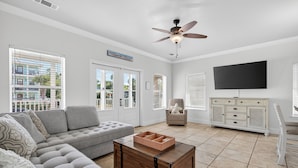 A living room with a grey sectional sofa, a wooden coffee table, a beige armchair, a TV, and a ceiling fan. - Destined to Be - Beach Cottage with Private Pool & Guest House (Destin)