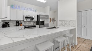 A modern kitchen with white cabinetry, a marble countertop, a stainless steel refrigerator, and a coffee maker. - Destined to Be - Beach Cottage with Private Pool & Guest House (Destin)