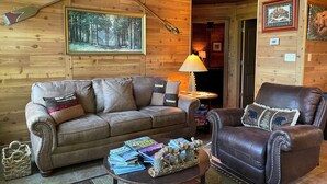 A cozy cabin interior with a wooden wall, a ceiling fan, a framed forest painting, a leather sofa, a leather armchair, and a small table with magazines and a decorative log. - Secluded yet within minutes of your favorite outdoor activities!! (Marshall)
