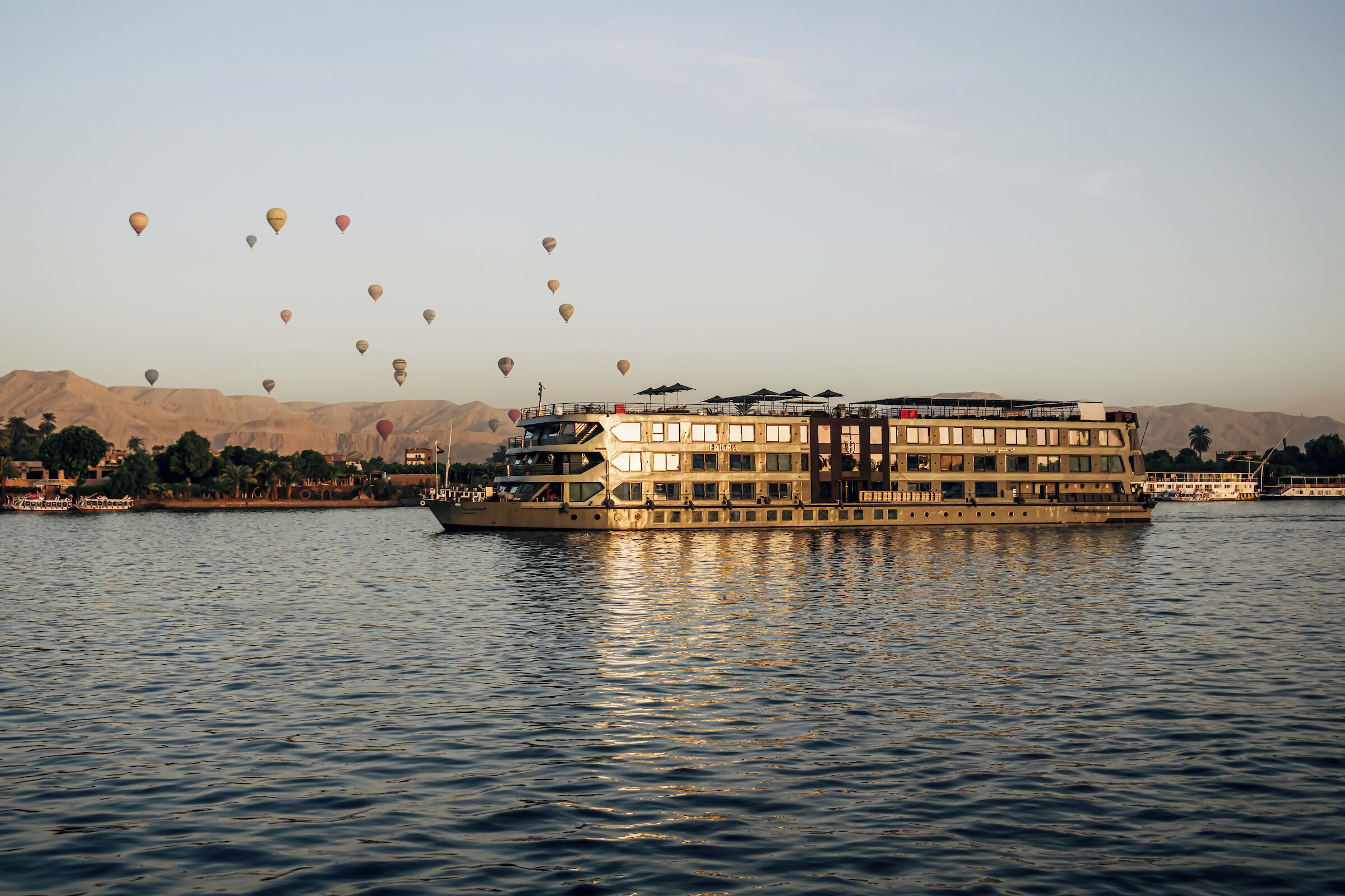 A large riverboat with multiple decks and a covered upper deck, floating on a river with a backdrop of mountains and a clear sky.