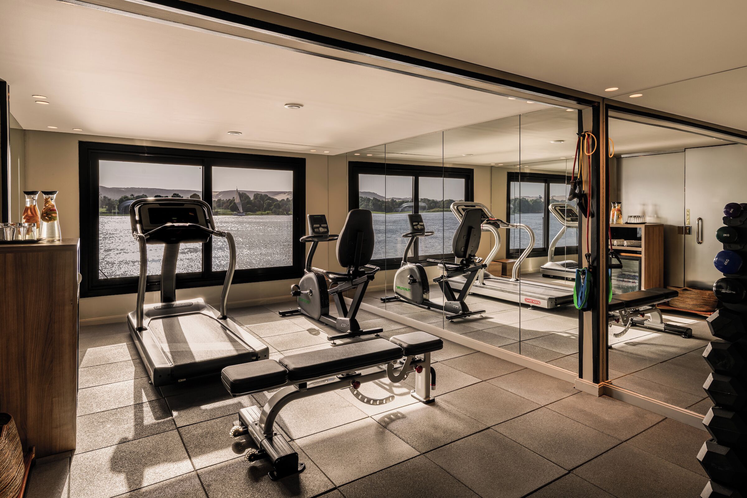 A gym with treadmills, elliptical machines, and a bench. It has large windows with a view of a body of water and hills.