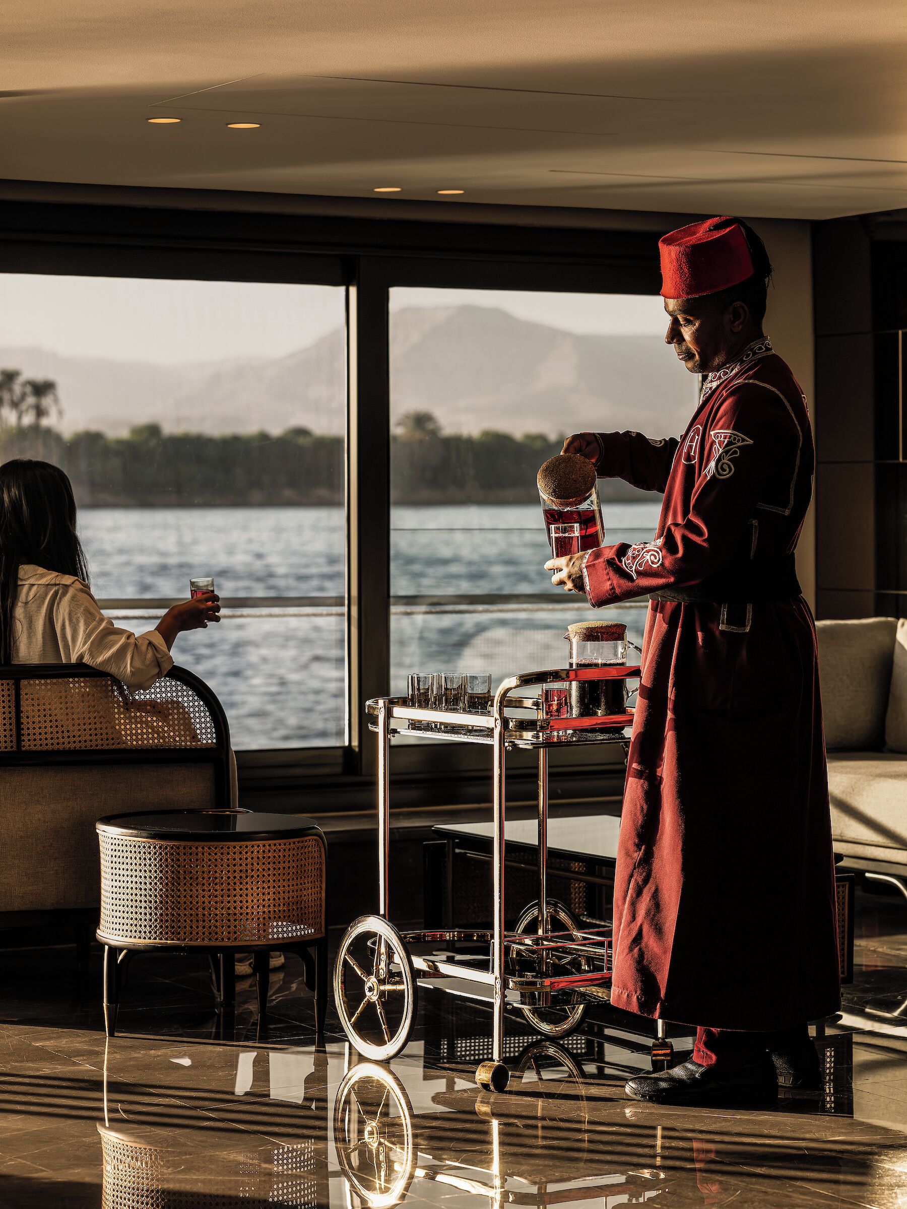 Una persona vestida con traje tradicional sirve bebidas en una mesa con vistas a un lago y montañas.