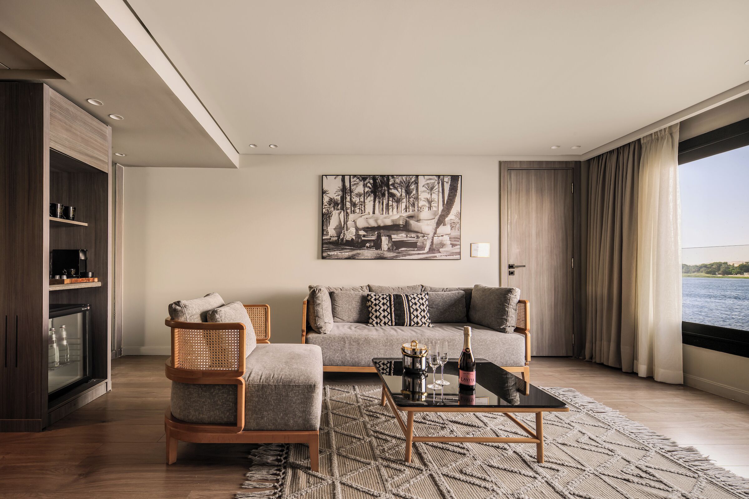 A modern living room with a sofa, armchair, and coffee table, a fireplace, and a large window with a view of the water.
