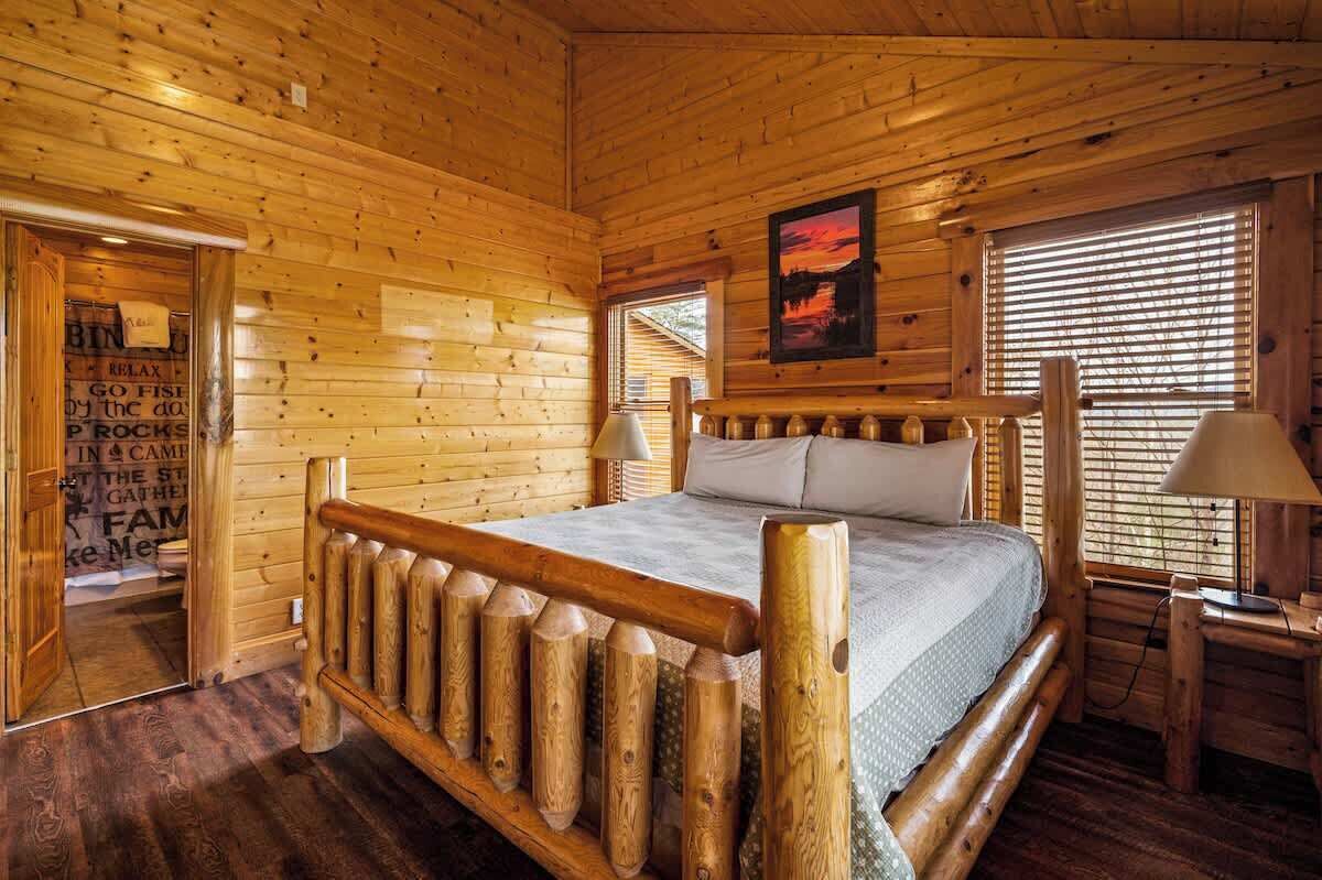A wooden cabin bedroom with a bed, a nightstand, a lamp, and a window with blinds.