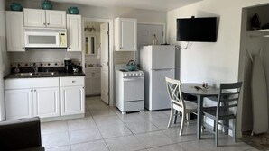 Eine Küche mit weißen Schränken, einem Herd, einer Mikrowelle und einem Kühlschrank. Ein Esstisch mit Stühlen und ein an der Wand befestigter Fernseher sind ebenfalls vorhanden.