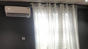 En soffa i mörk färg med två gula kuddar, en vit luftkonditioneringsenhet på väggen och ett fönster med tunna, genomskinliga gardiner.