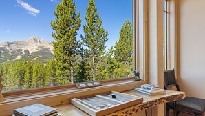 A room with a view of mountains, a window with a wooden desk, and a chair.