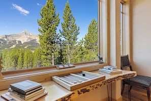A room with a view of mountains, a window with a wooden desk, and a chair.