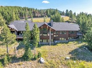 A two-story log cabin with a deck, surrounded by forest and hills.