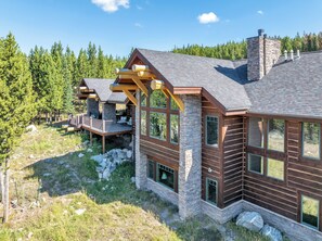 A log cabin with a stone foundation, large windows, and a deck area. - White Wolf Retreat @ Big Sky Resort | Sleeps up to 21! (Big Sky)