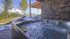 A hot tub with a stone wall backdrop, surrounded by snow and trees, with a mountain in the background.