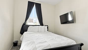 A single bed with a black headboard, a flat-screen TV mounted on the wall, and a window with black curtains.