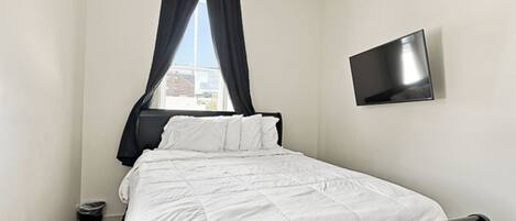 Een enkel bed met een zwart hoofdeinde, een flatscreen televisie aan de muur en een raam met donkere gordijnen.