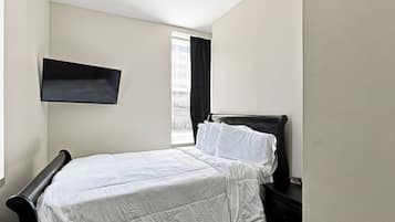 A bedroom with a bed, a nightstand, a wall-mounted shelf, and a window with curtains.