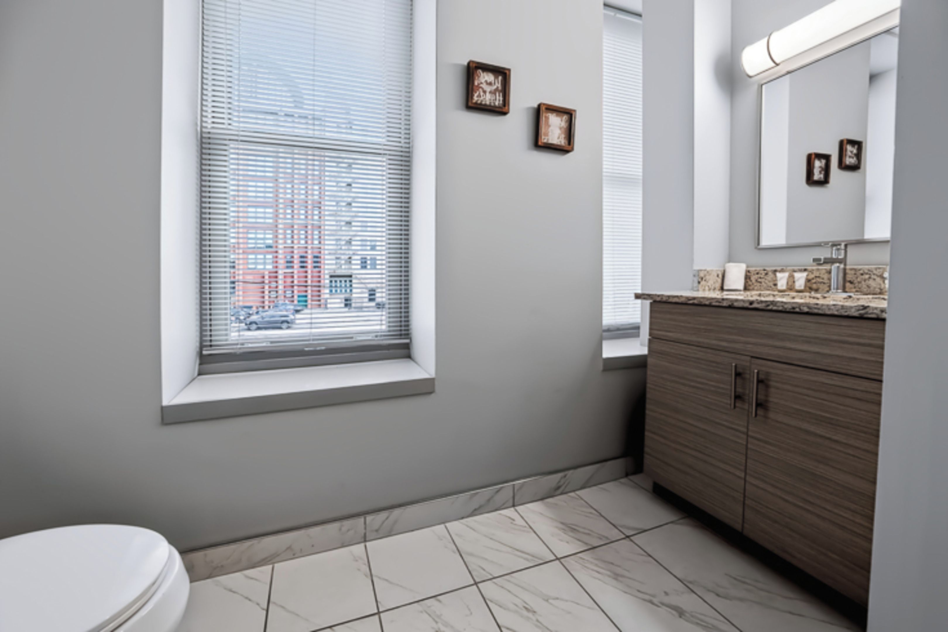 Une salle de bain comprenant des toilettes, une fenêtre avec des stores, un meuble-lavabo avec un miroir et un sol carrelé.